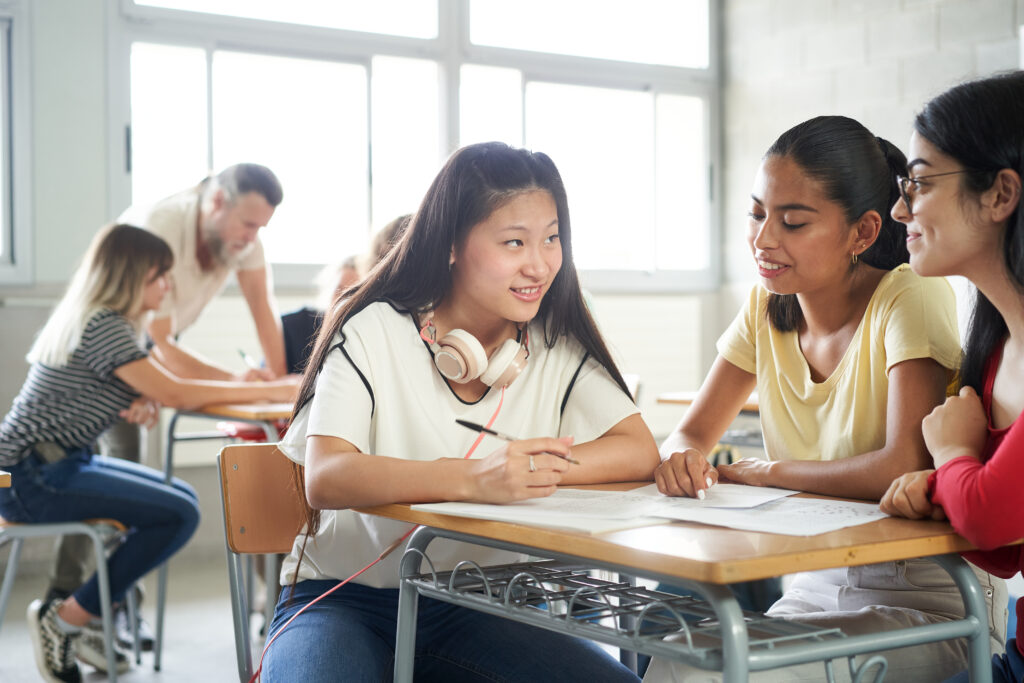 Smiling students working together as a team in class in the background the teacher helps other classmates